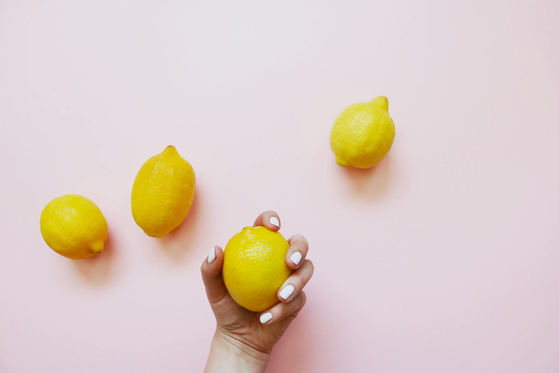 A hand with white nails holding a lemon on a soft pink background, surrounded by lemons.
